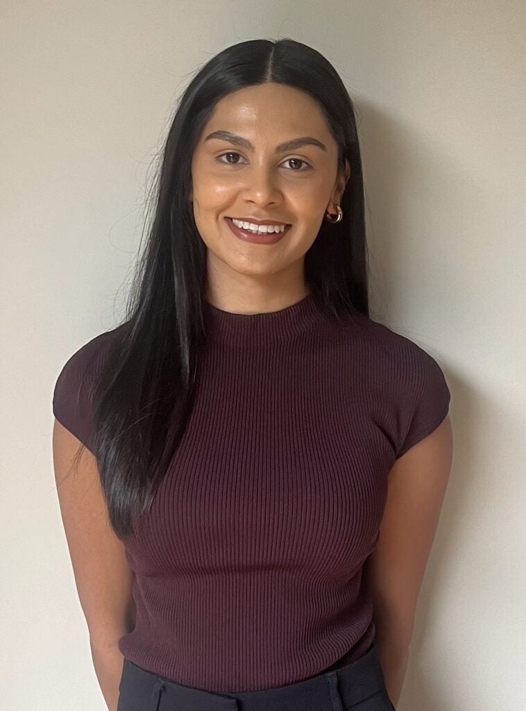 A woman with long dark hair smiles while standing against a plain light-coloured wall. She is wearing a ribbed, short-sleeved maroon top and small gold hoop earrings.