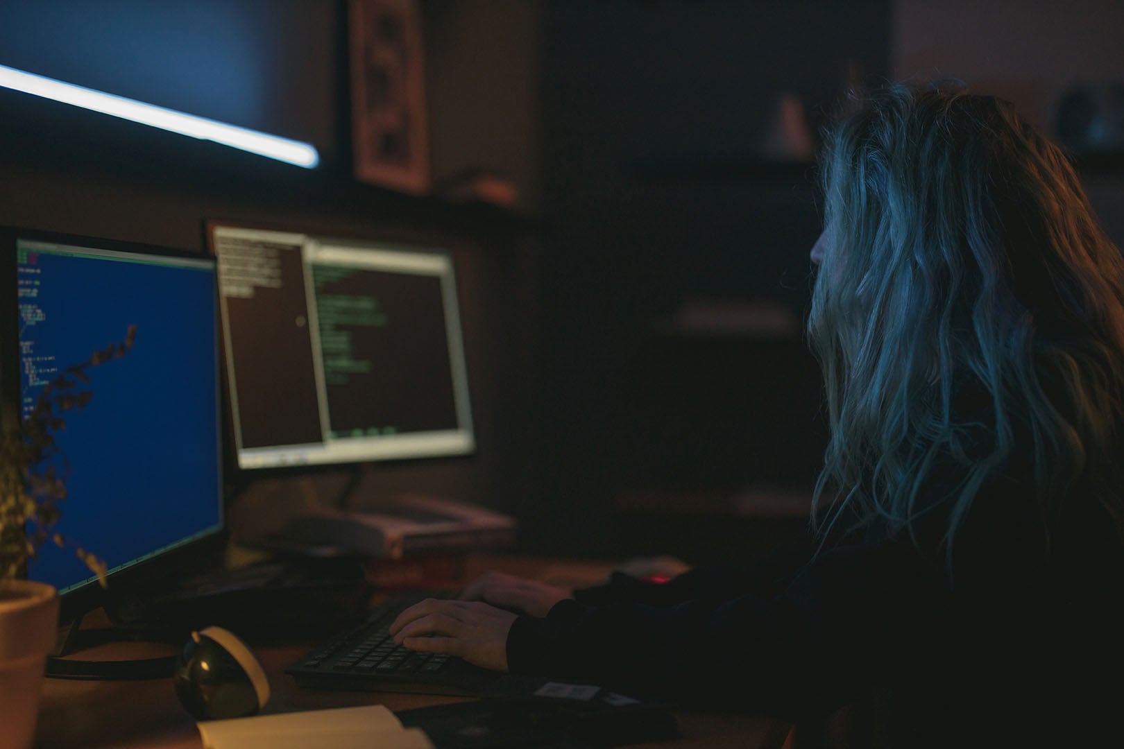 Woman with blonde hair and wearing a black jumper working in cyber security in a dark office. She'd typing on a keyword and looking at two computer screens with code on it.