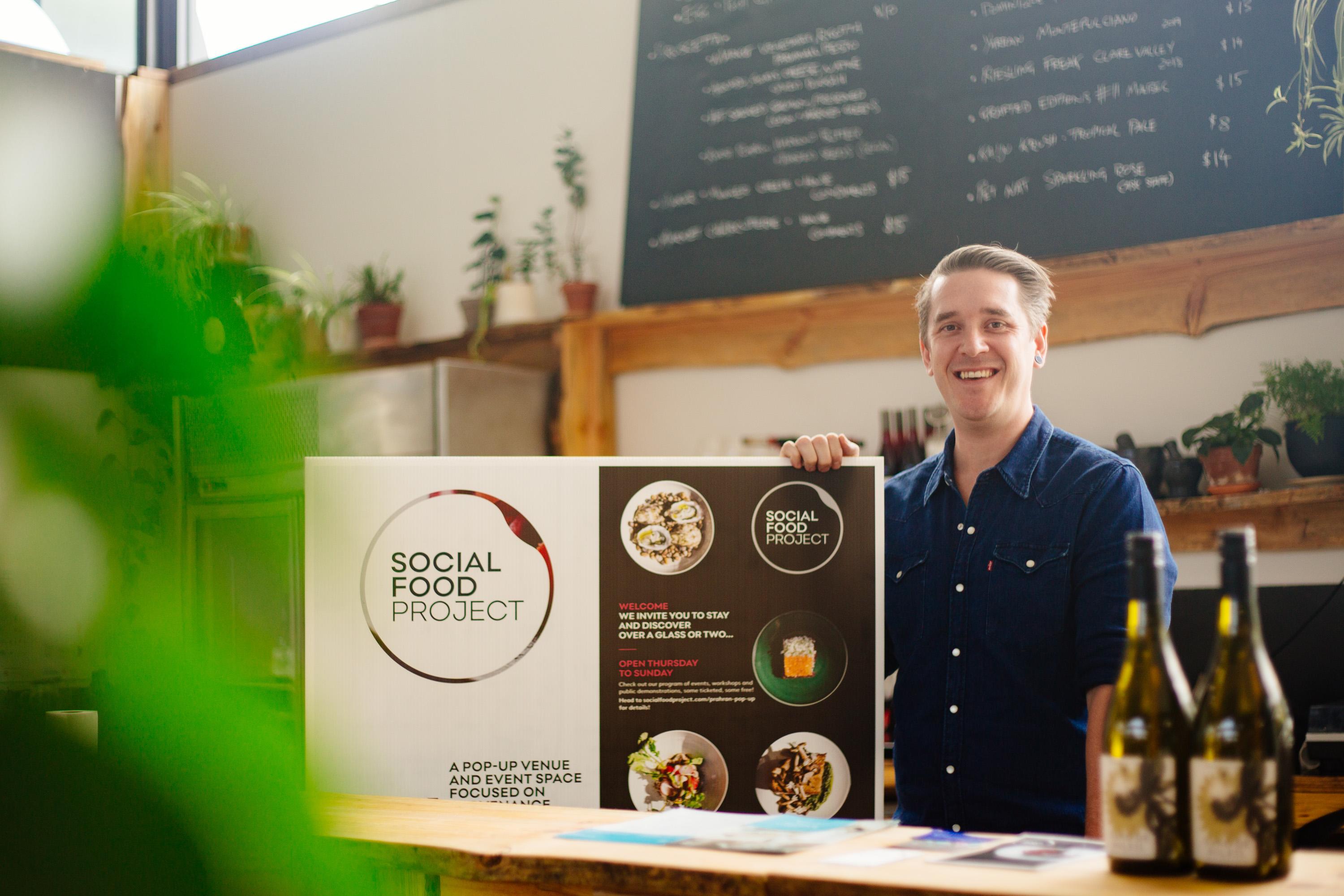 Ben McMenamin at the Social Food Project pop-up in Prahran, holding the menu sign.